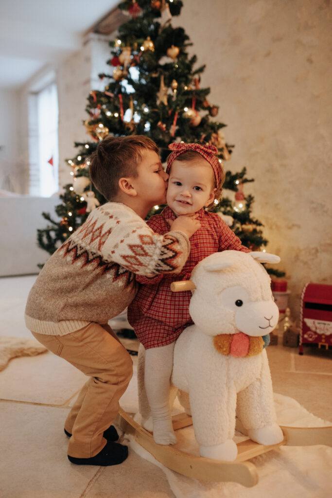 Bisou entre frère et sœur devant le sapin, séance photo de Noël