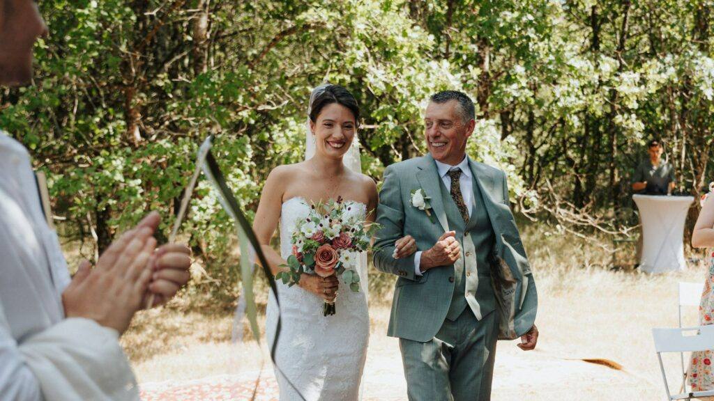 Entrée de la mariée souriante au bras de son papa, cérémonie laïque au Domaine de la Volière