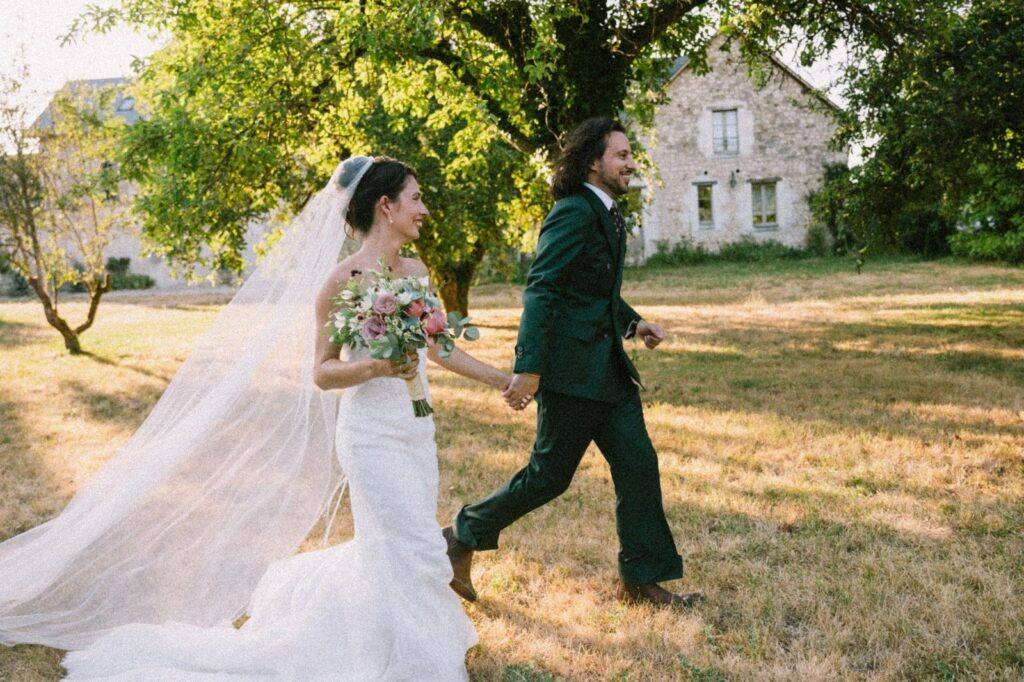 Les mariés se promènent dans le parc devant une maison en pierre du Domaine de la Volière