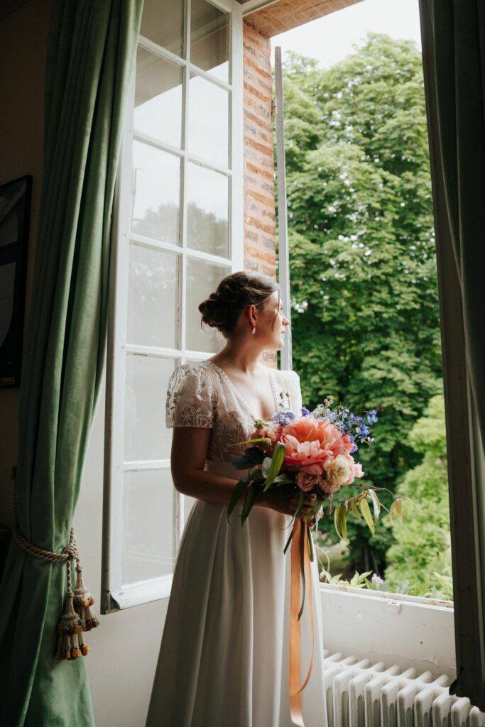 La mariée avec son bouquet près d'une fenêtre pendant les préparatifs au Manoir du Grand Courtoiseau